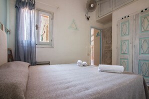 Ein Schlafzimmer mit einem großen Bett, einem Fenster mit Vorhängen, einem Ventilator und einer Tür mit geometrischen Mustern.