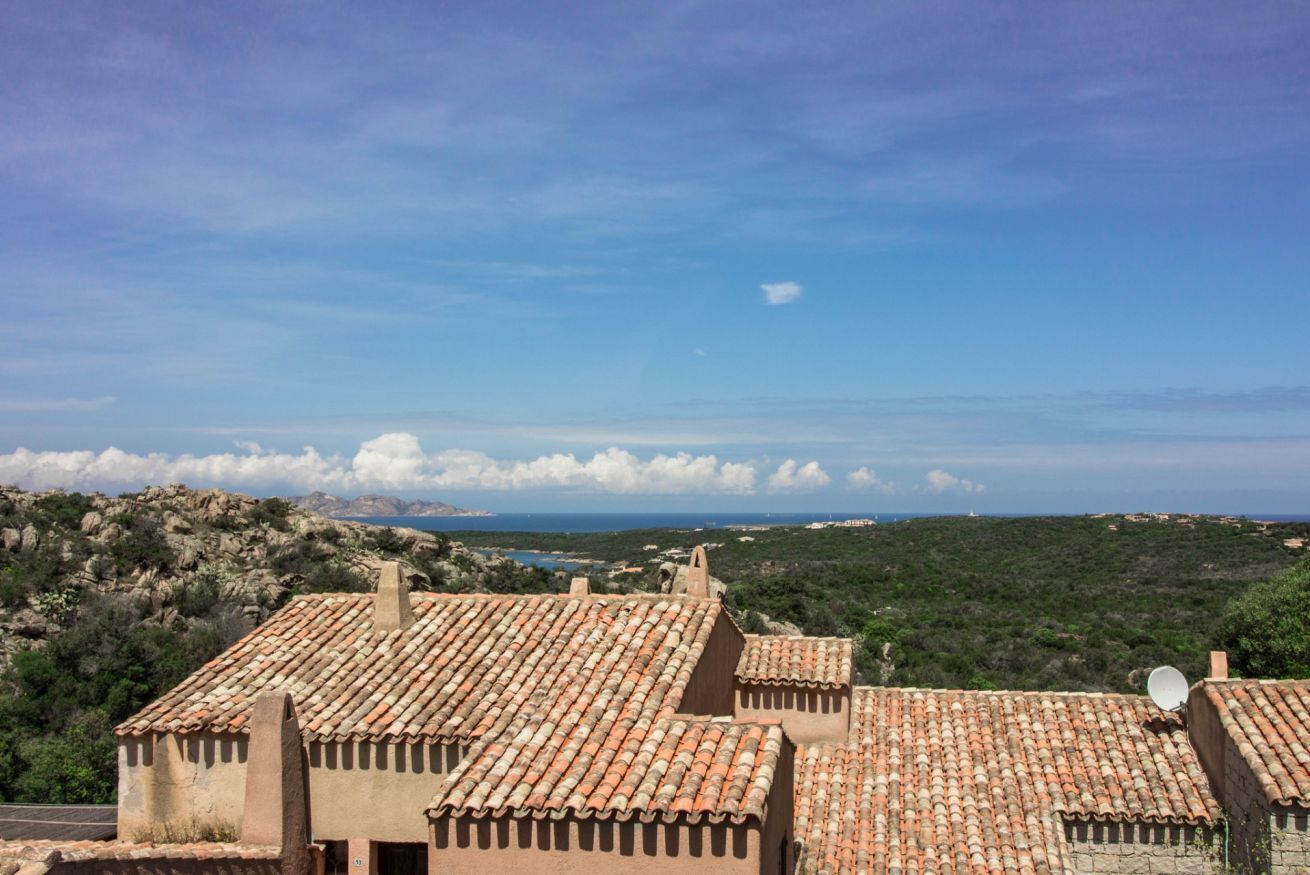 A coastal village with terracotta-roofed buildings, a clear sky, and a distant view of the sea.
