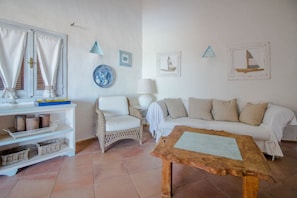 A living room with a white sofa, a wicker chair, a wooden coffee table, a white console table with shelves, and decorative wall art. - Cozy and bright 80 square meters apartment, perfect for four people, located on the first floor of a condominium with a swimming pool (no elevator).The property is located in Liscia di Vacca, a short distance from Porto Cervo, reachable by car or public  (Liscia di Vacca)