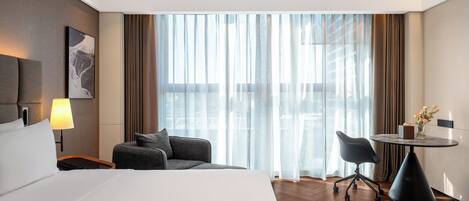 A hotel room with a large bed, a desk, a chair, a lamp, and a window with curtains.