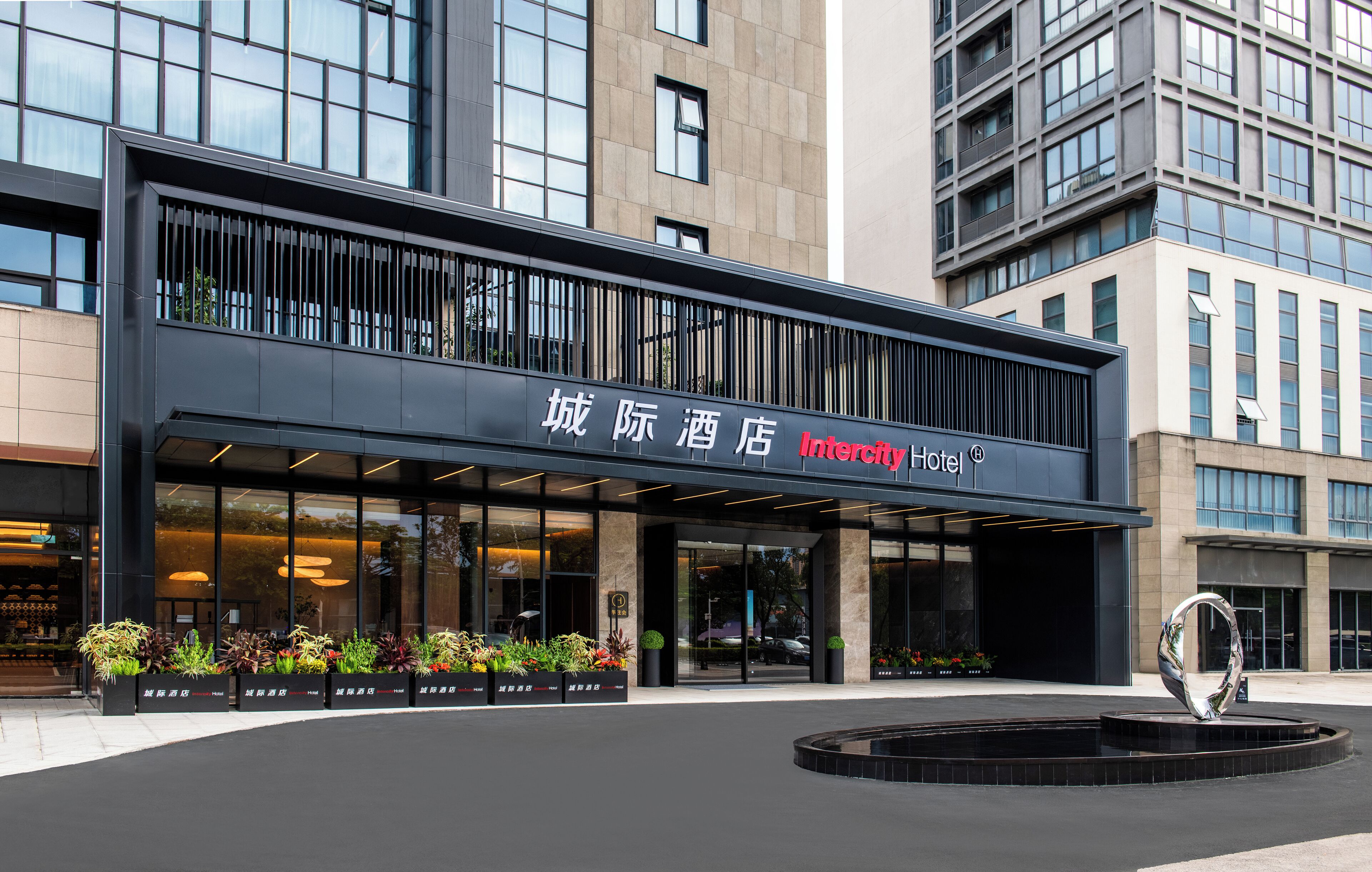 A modern hotel entrance with a large sign, glass doors, and a decorative metal sculpture in front.