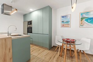 Una cuina moderna amb forn integrat, un rentaplat i zona de menjador amb taula de vidre i cadires blanques.