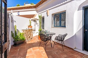 Un pati amb sòl de rajoles, una taula amb cadires, plantes en testos i una porta amb un senyal amb la lletra 'C'.
