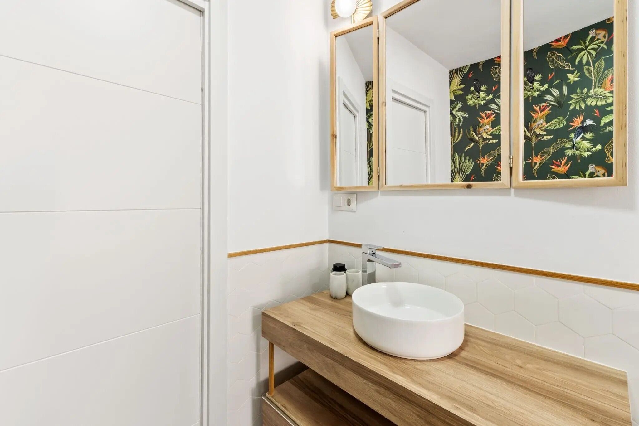 Een badkamer met witte tegels, een houten wastafelmeubel, een rond wastafel en een spiegel met bloemenbehang.
