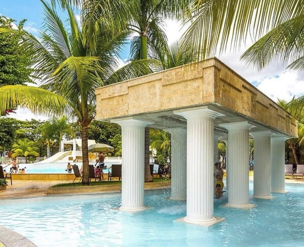A pool area with a covered seating area supported by columns, surrounded by palm trees. - HOTEL LACQUA DI ROMA  (Caldas Novas)