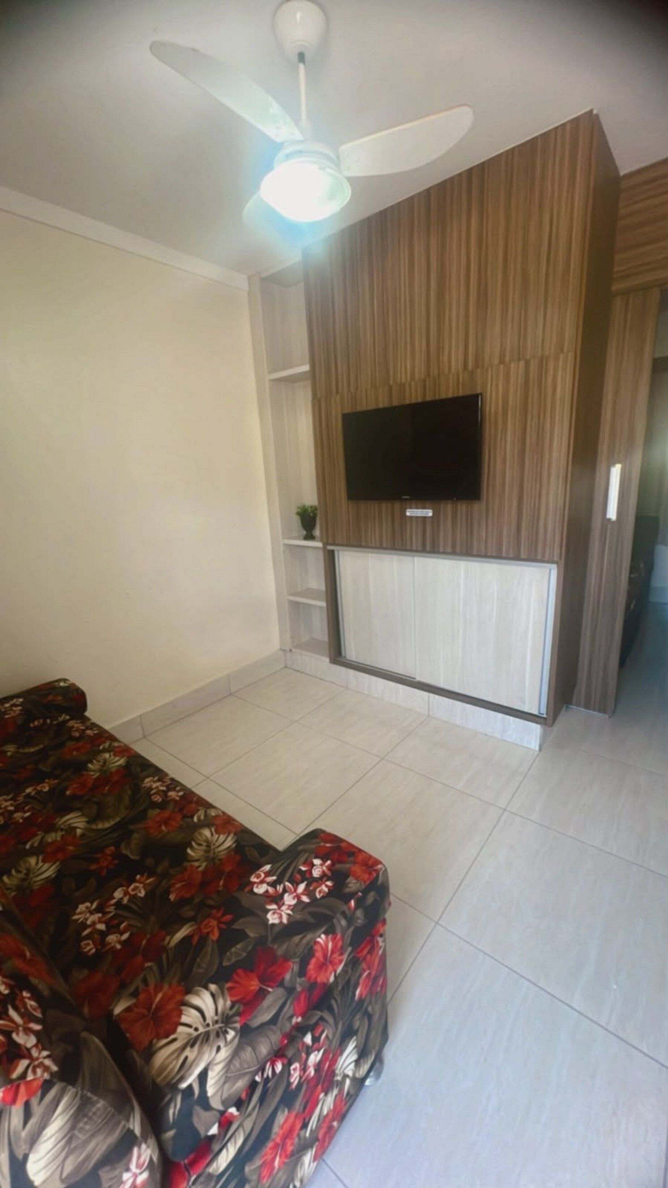 A bedroom with a bed, a ceiling fan, a flat-screen TV mounted on a wooden panel wall, and a tiled floor.
