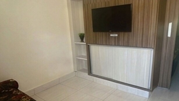 A room with a flat-screen TV mounted on a wooden panel wall, a white door, and a tiled floor. - HOTEL LACQUA DI ROMA  (Caldas Novas)