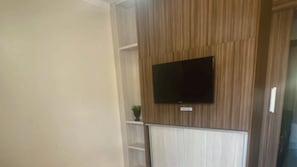 Quarto com televisão de tela plana fixada numa parede de madeira, radiador branco embaixo e uma pequena planta em vaso sobre uma prateleira.