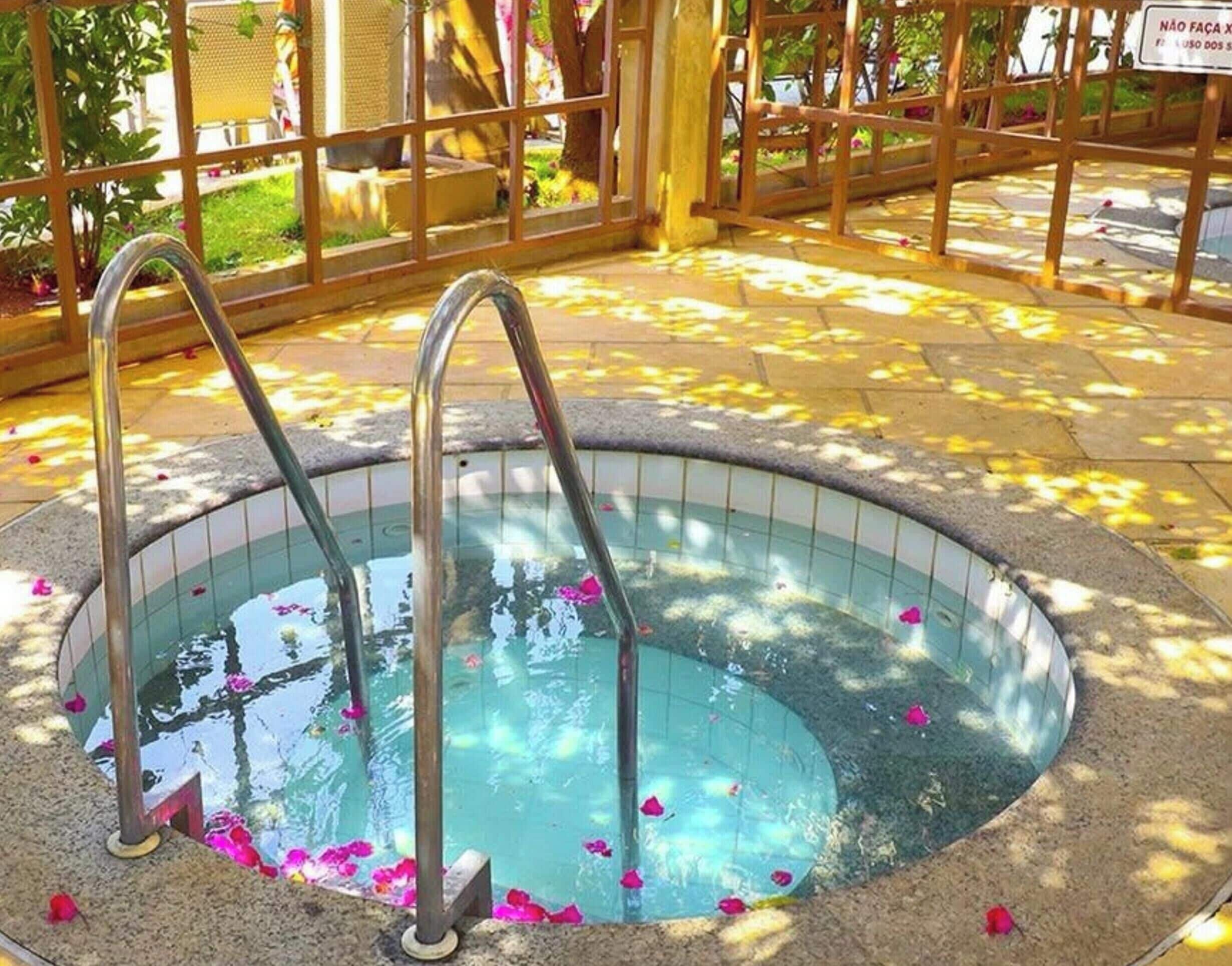 Un bain à remous avec des marches en métal et un bord carrelé, entouré d’une clôture en bois et d’une zone pavée parsemée de pétales de fleurs.