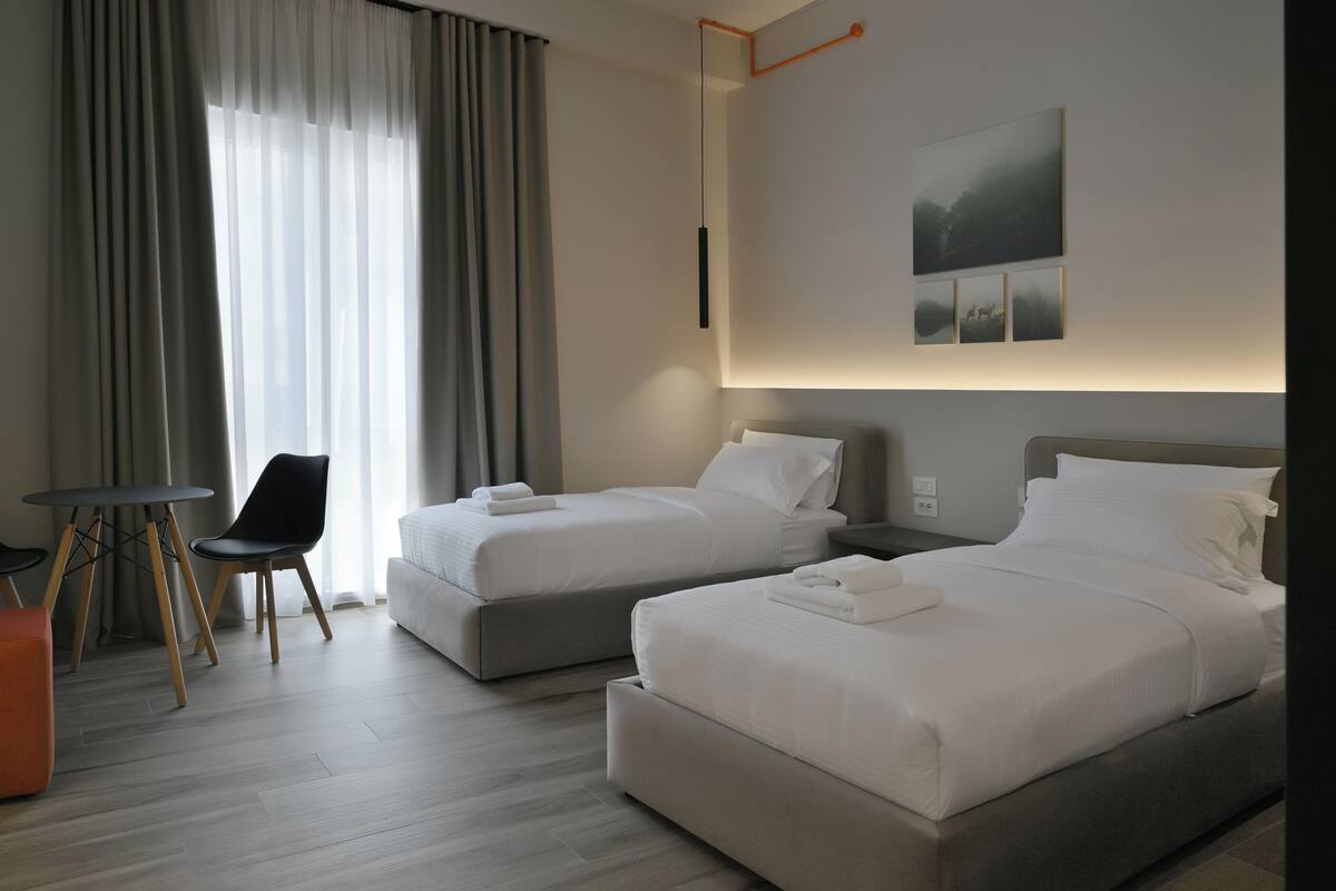 A modern hotel room with two beds, a small table, and a chair.