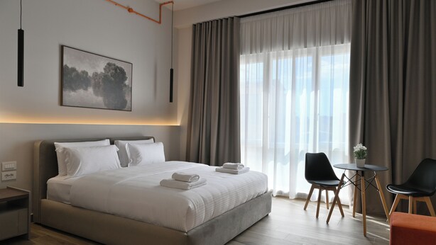 A modern hotel room with a large bed, a small table, two chairs, and a view of the city through sheer curtains. - Cosmos Luxury Studio Suites (Tirana)