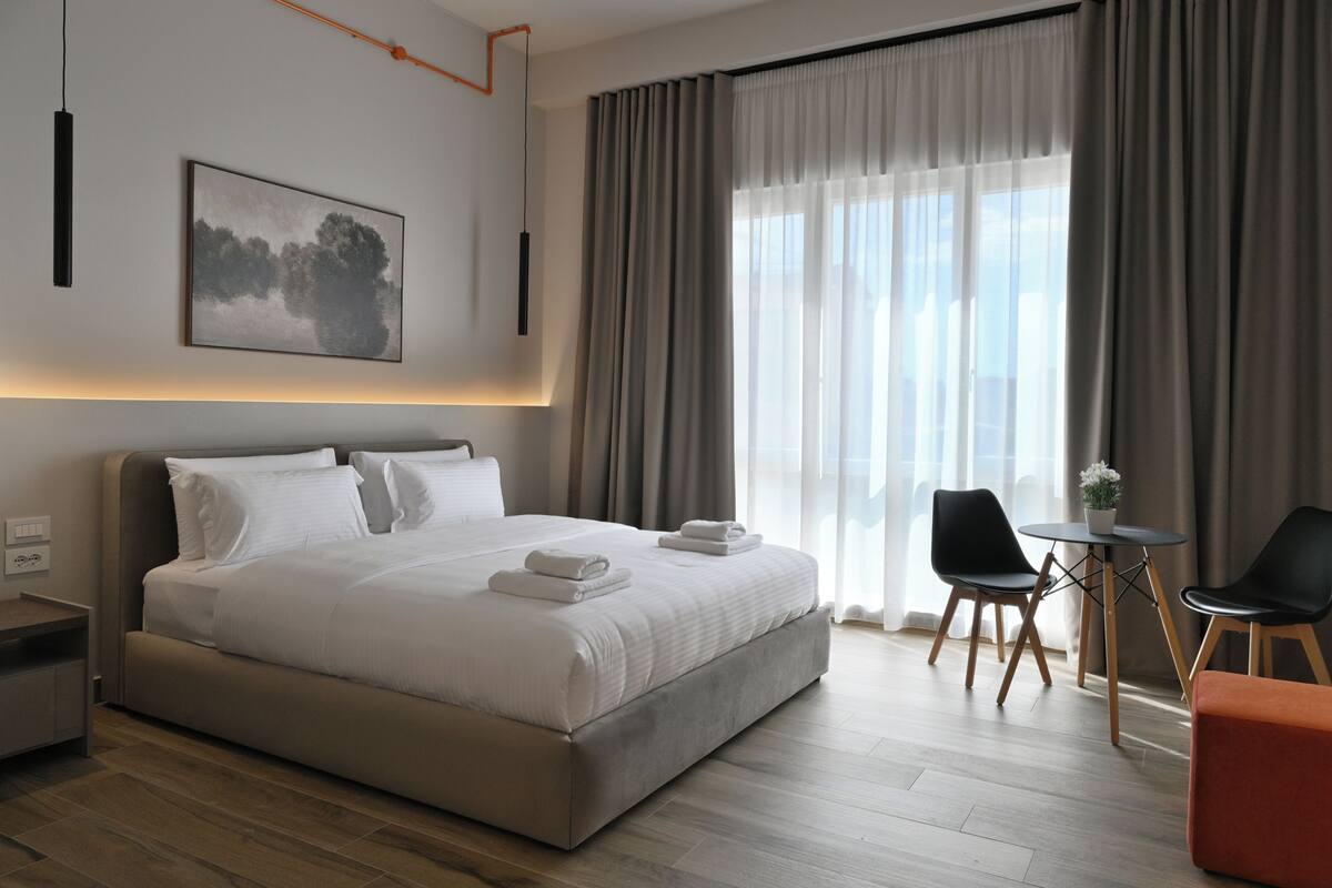 A modern hotel room with a large bed, a small table, two chairs, and a view of the city through sheer curtains.