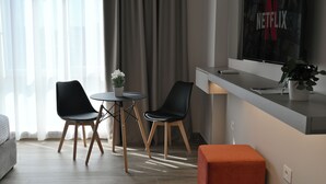 A modern hotel room with a bed, a desk with a laptop, two chairs, a small table with a plant, and a large window with curtains. - Cosmos Luxury Studio Suites (Tirana)