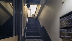 Una escalera dentro de un edificio con barandilla, un estante en la pared y una puerta al final.