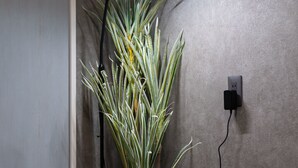 Una planta en maceta junto a una lámpara de pie en una esquina, con una pared gris e un enchufe.