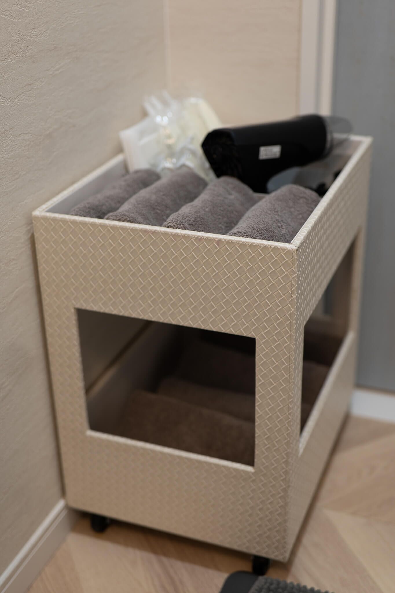 Un pequeño mueble de almacenamiento beige con textura y una abertura central, que contiene toallas enrolladas y un objeto cilíndrico negro.