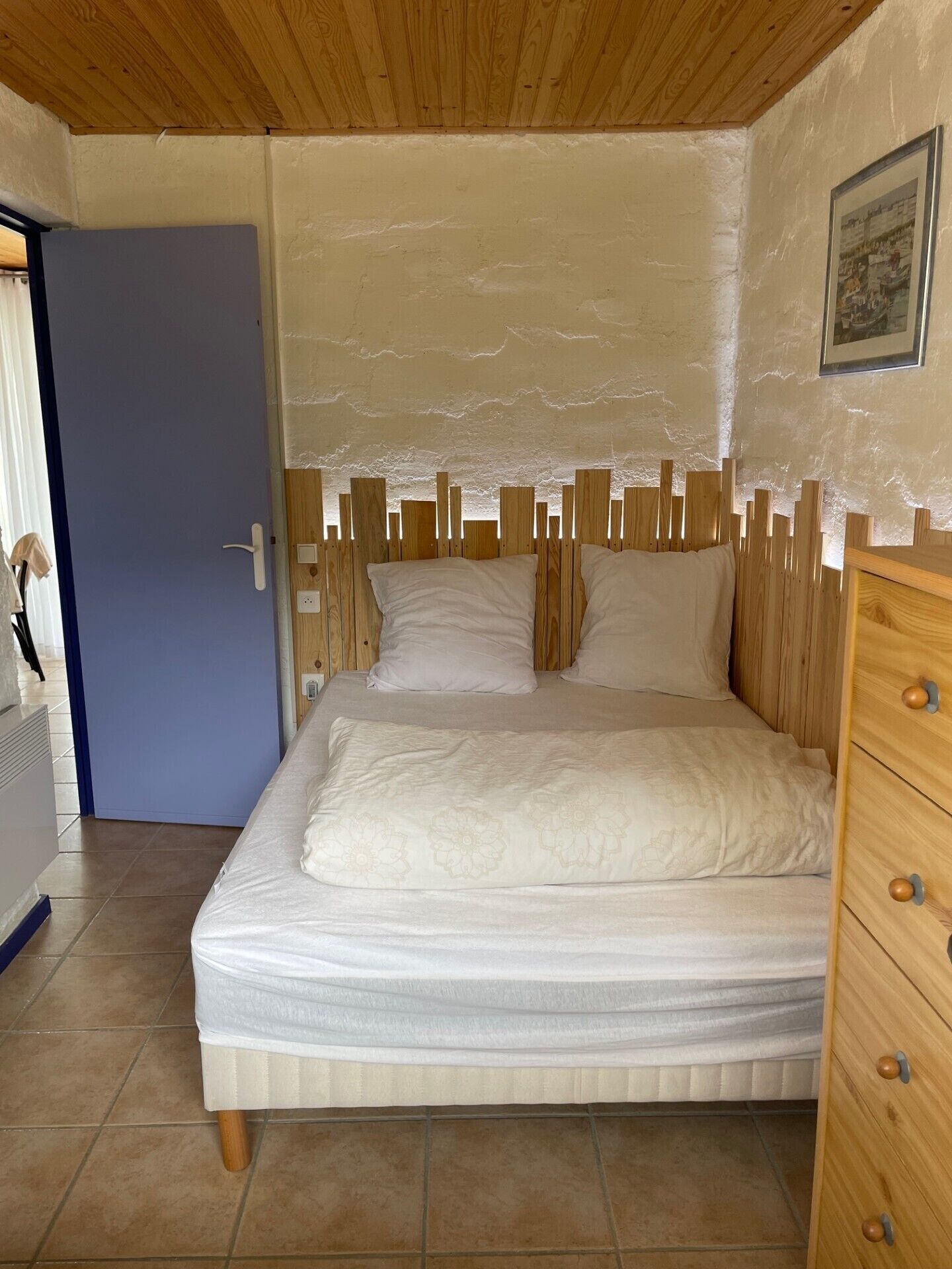 Une chambre à coucher avec une tête de lit en bois, un lit recouvert de draps blancs, une commode en bois dotée de tiroirs et une porte donnant sur une autre pièce.