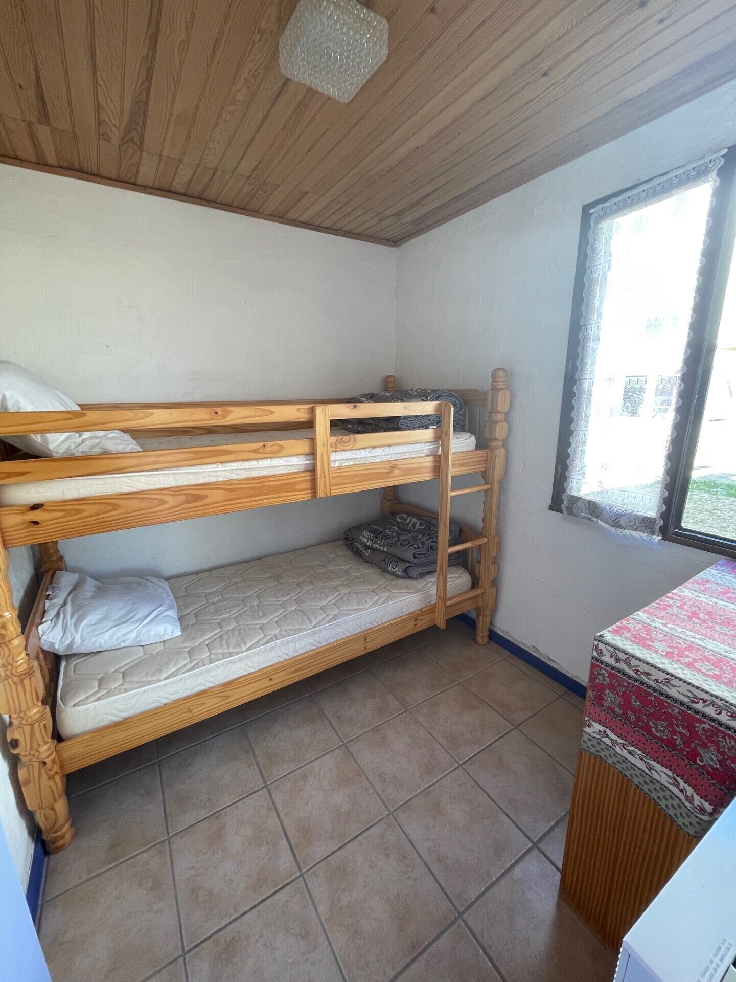 A bunk bed room with a window, a small table with a patterned tablecloth, and a tiled floor.