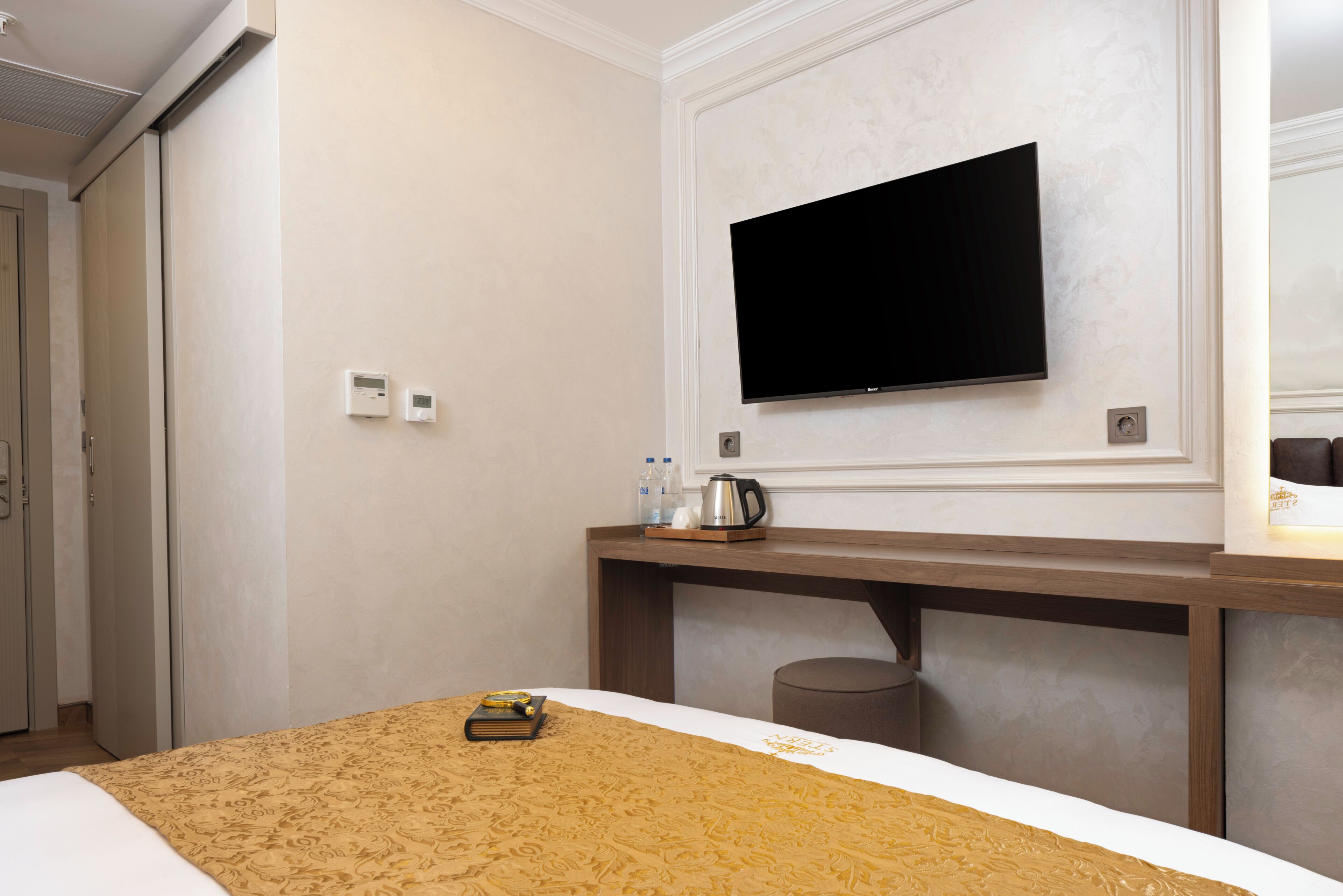 Ein Hotelzimmer mit einem Bett, einem flachbildfernseher an der Wand, einem Nachttisch mit Wasserkocher und Flaschen sowie einer kleinen Mülltonne.