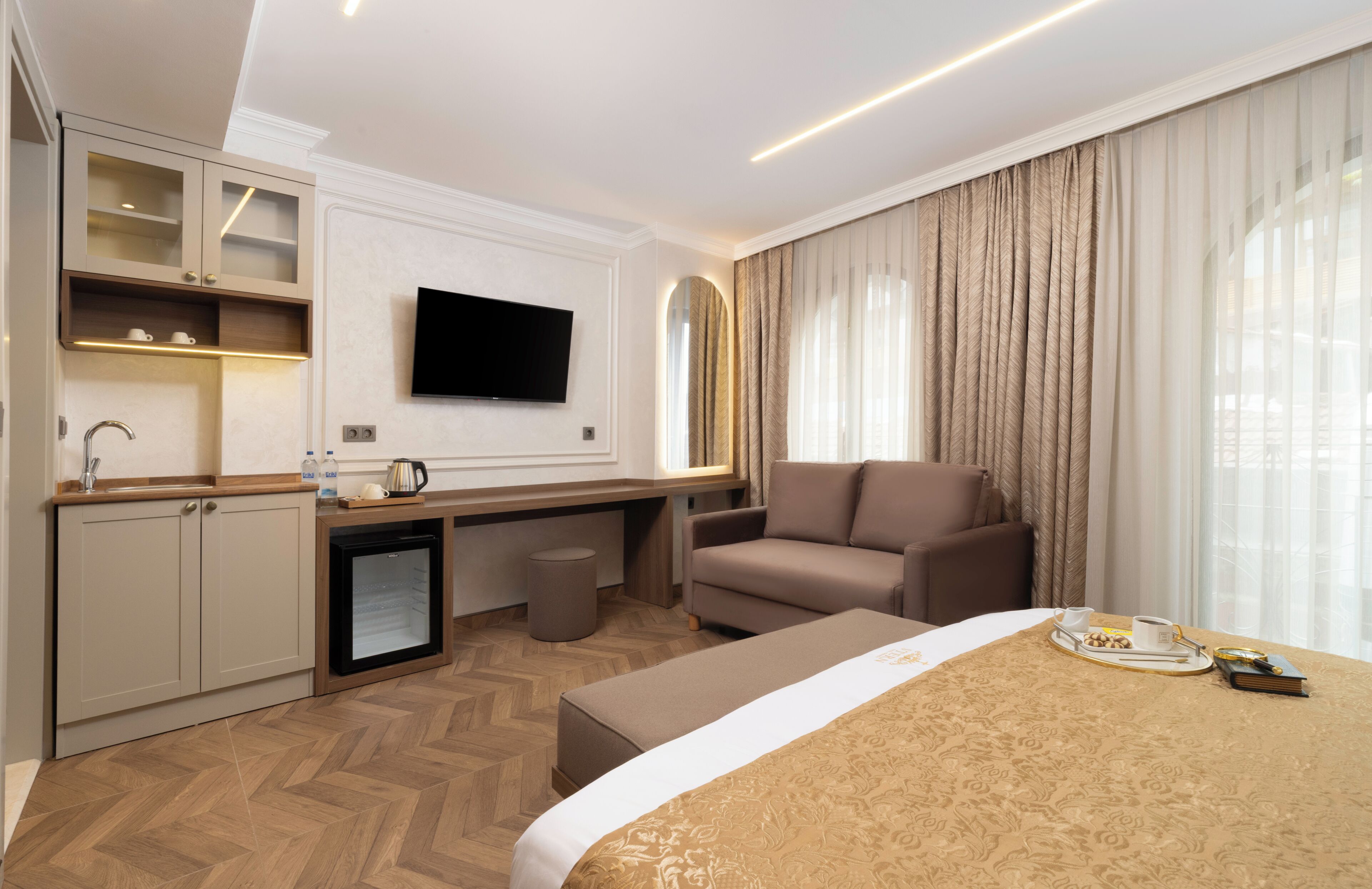 Ein Hotelzimmer mit einem Bett, einem Sofa, einem Schreibtisch mit Fernseher, einer Minibar und einem Fenster mit Vorhängen.