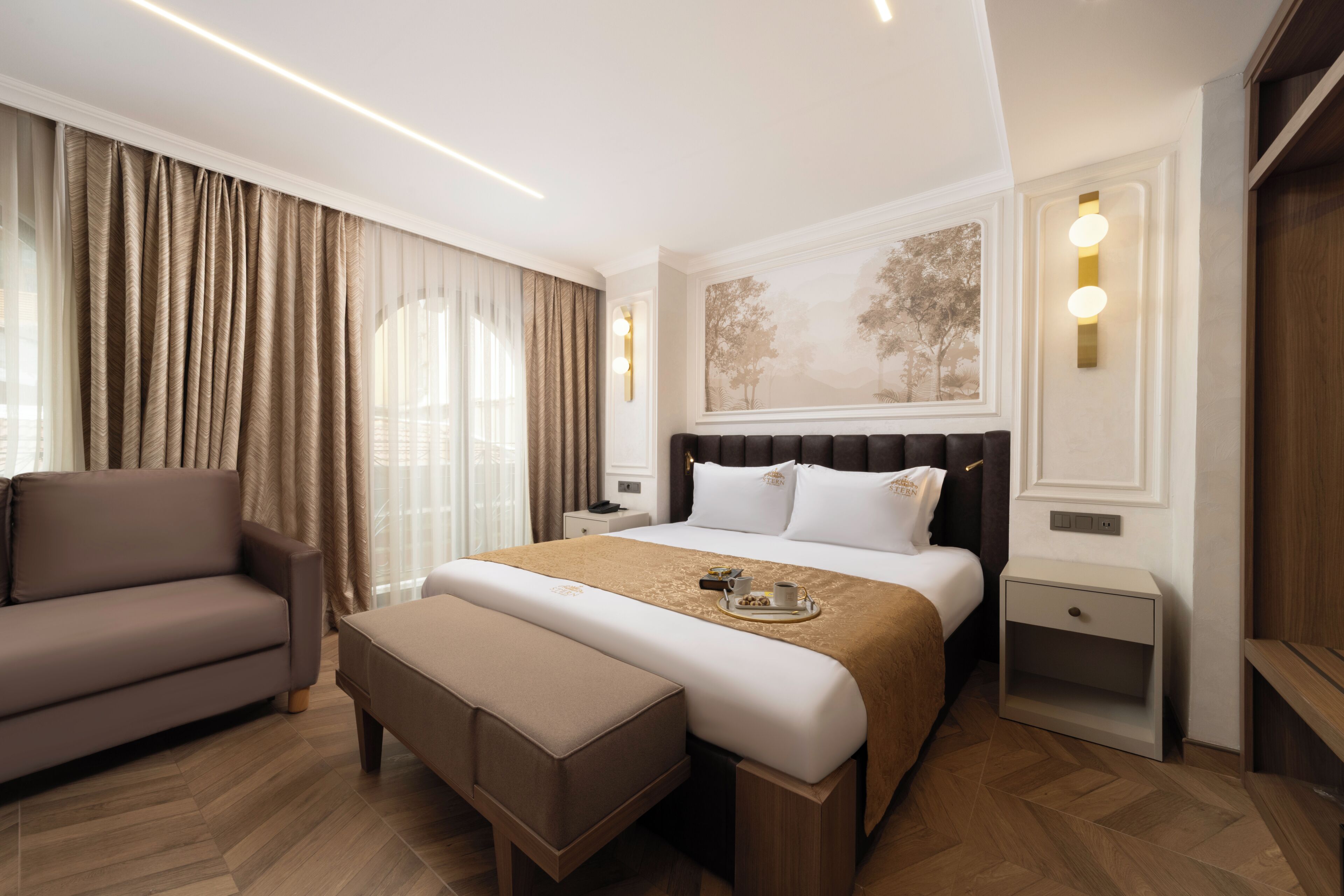 A hotel room with a bed, a sofa, a bench, a nightstand, and a wall-mounted light fixture.