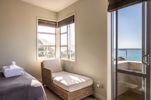 Ein Zimmer mit einem Bett, einem Korbstuhl, einem Balkon mit Meerblick und einem Fenster mit Jalousien.