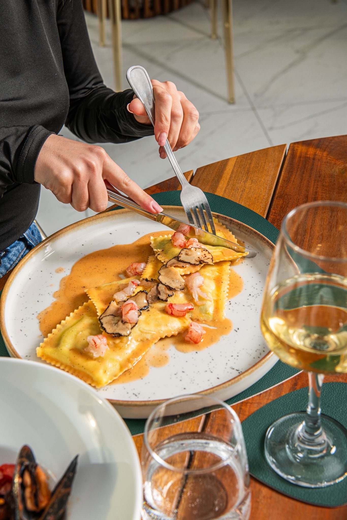 A person dining at a table with a plate of ravioli, a fork, and a glass of white wine.