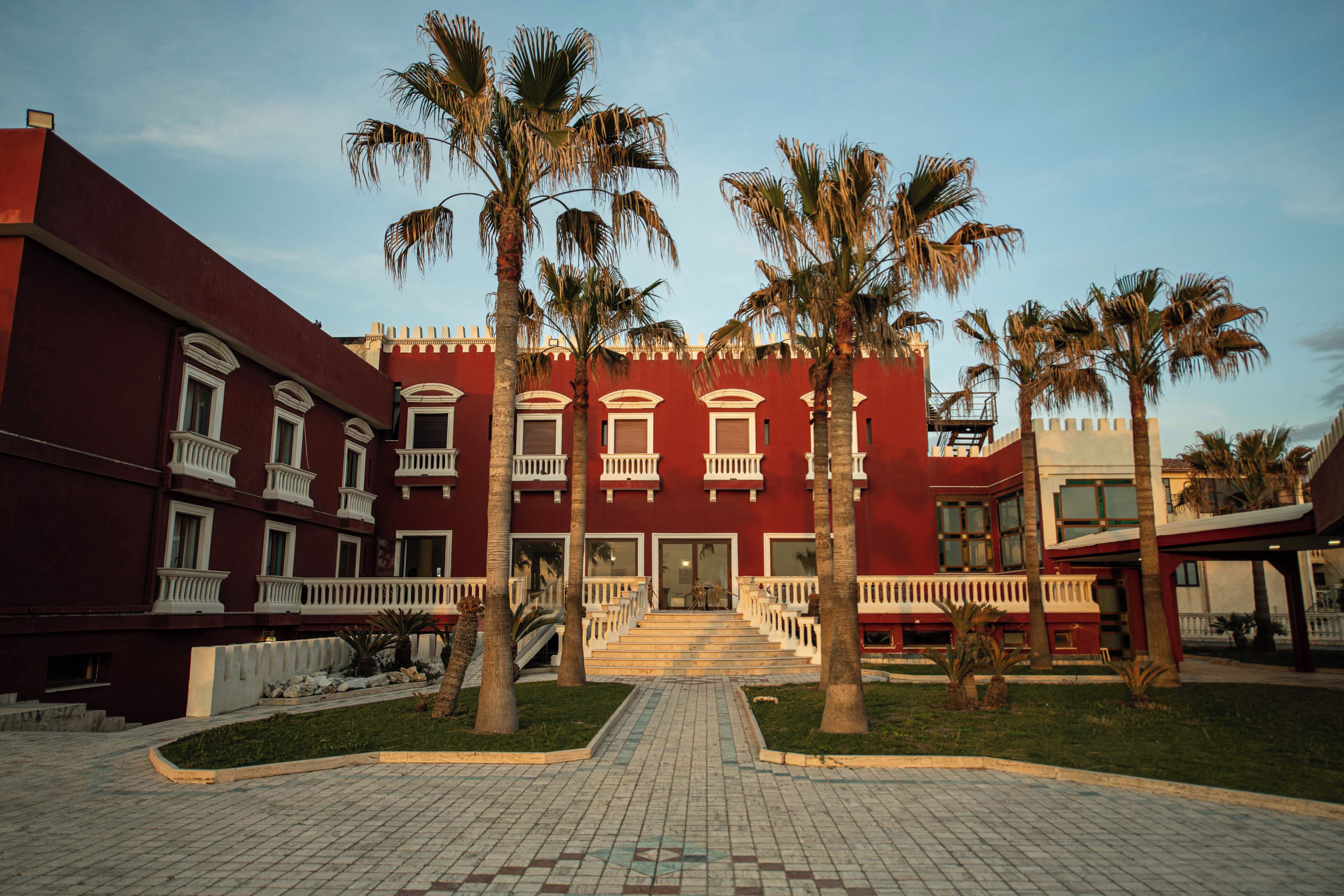 A red building with white balconies and palm trees in front.