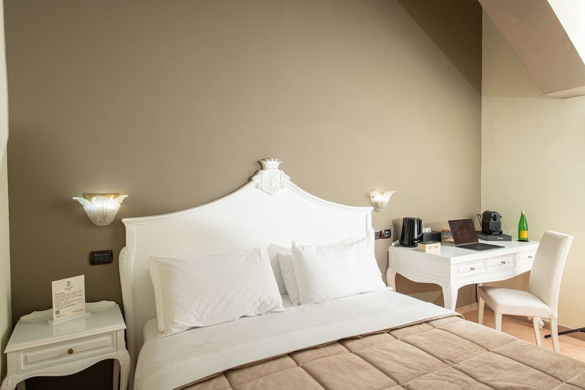 A bedroom with a white bed, a desk, and a chair. There is a nightstand with a lamp and a sign.