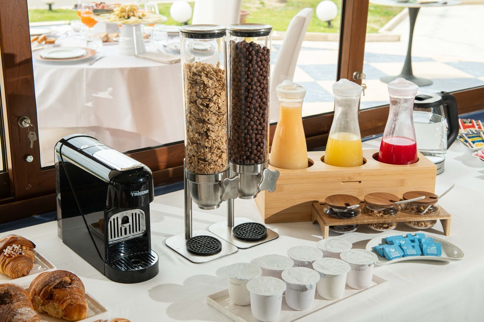 A breakfast buffet setup with a coffee machine, cereal dispensers, juice pitchers, and pastries.