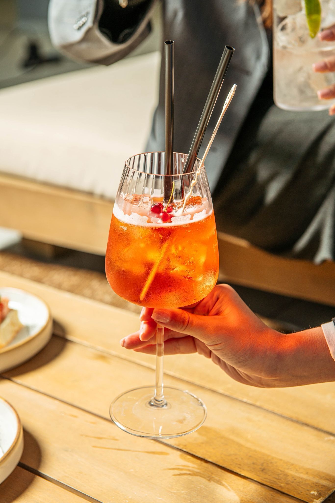 Eine Hand hält ein Cocktailglas mit Trinkhalm und einem Halter für Metallstrohhalme, Eiswürfeln und einem Limettenschnitz.