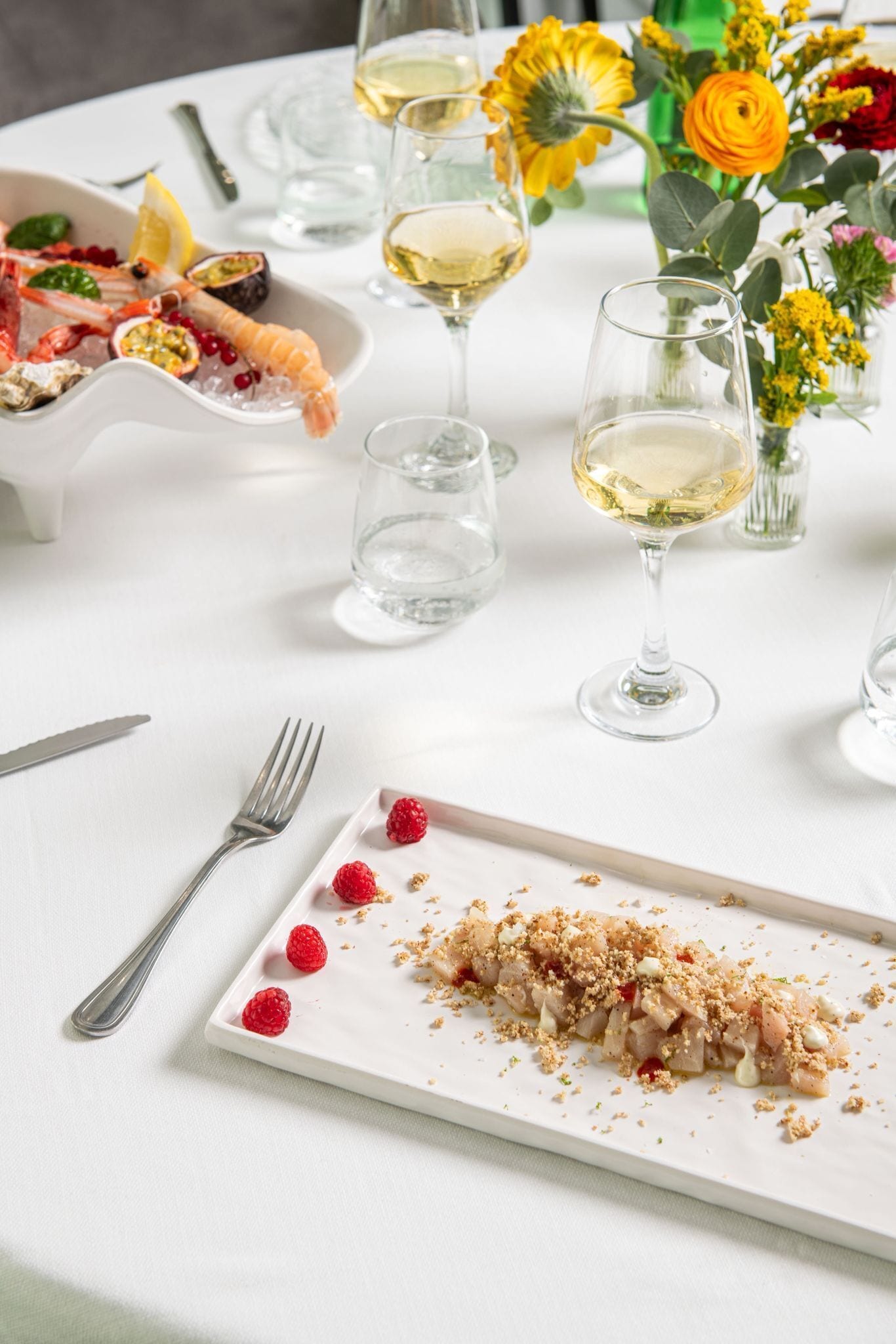 A dining table with a white tablecloth, a plate of food, a fork, a glass of white wine, and a vase with flowers.