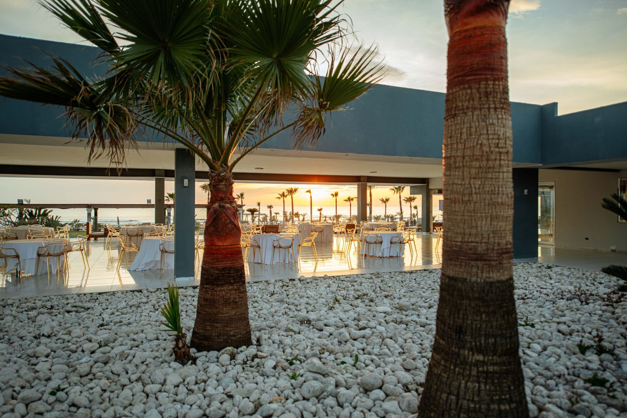 A resort with outdoor seating, palm trees, and a view of the sunset.