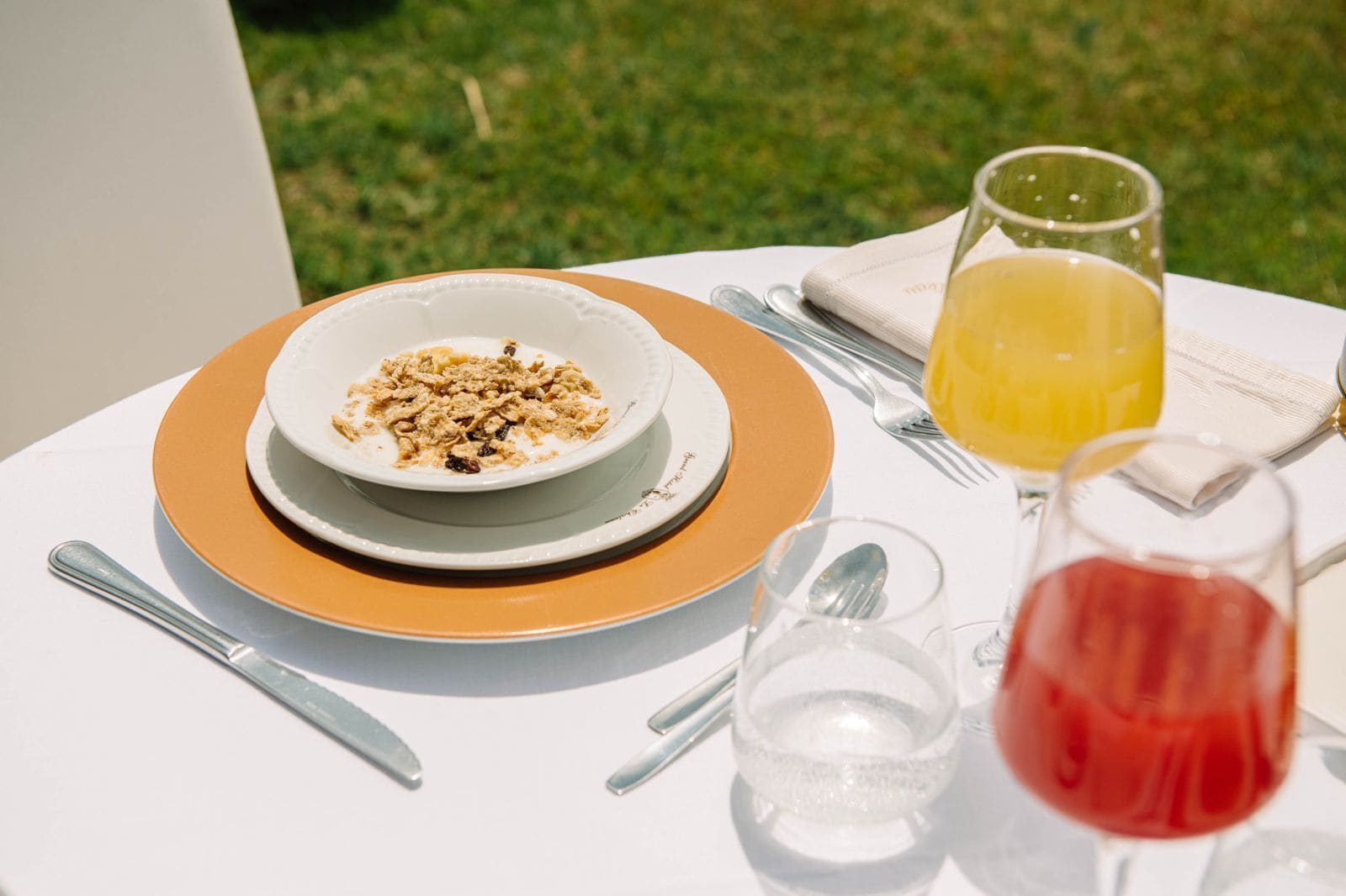 A table with a bowl of cereal, a glass of orange juice, a glass of red drink, and cutlery.