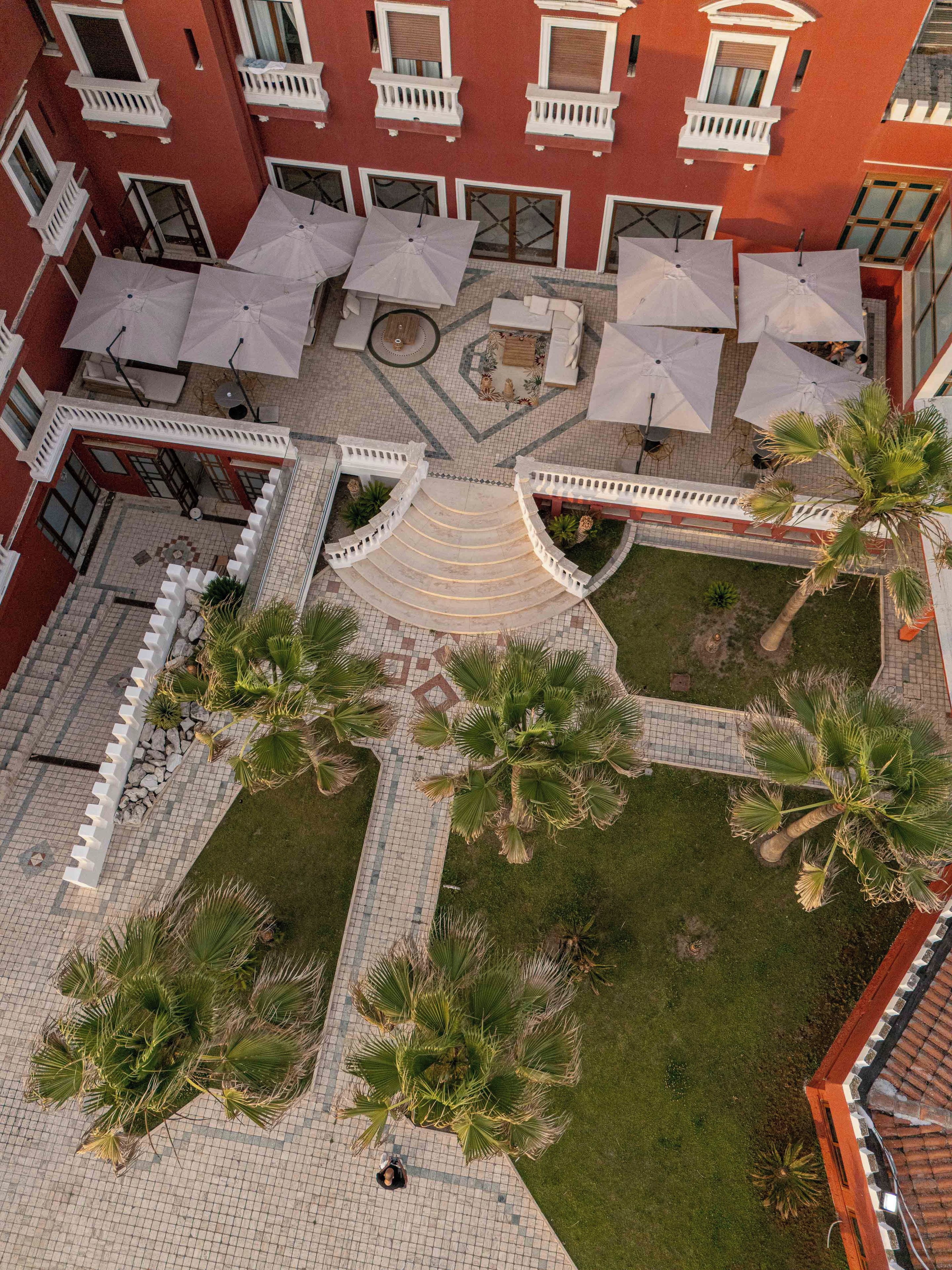 An aerial view of a courtyard with a central staircase, surrounded by palm trees and outdoor seating areas with umbrellas.