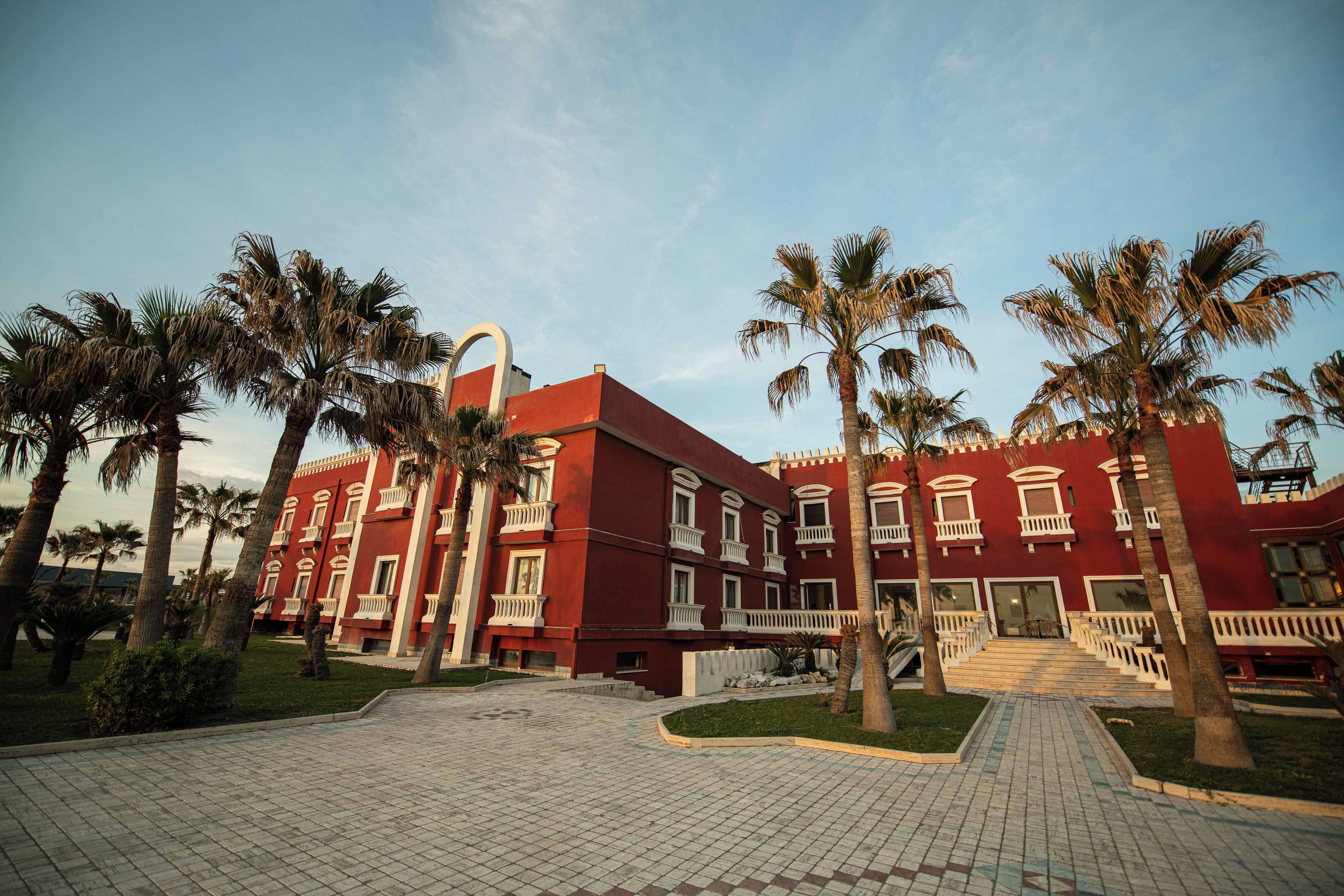 A red building with multiple balconies and palm trees in the foreground.