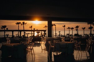 Un área de comedor al aire libre con mesas y sillas, con vistas a un atardecer con palmeras y un muelle.