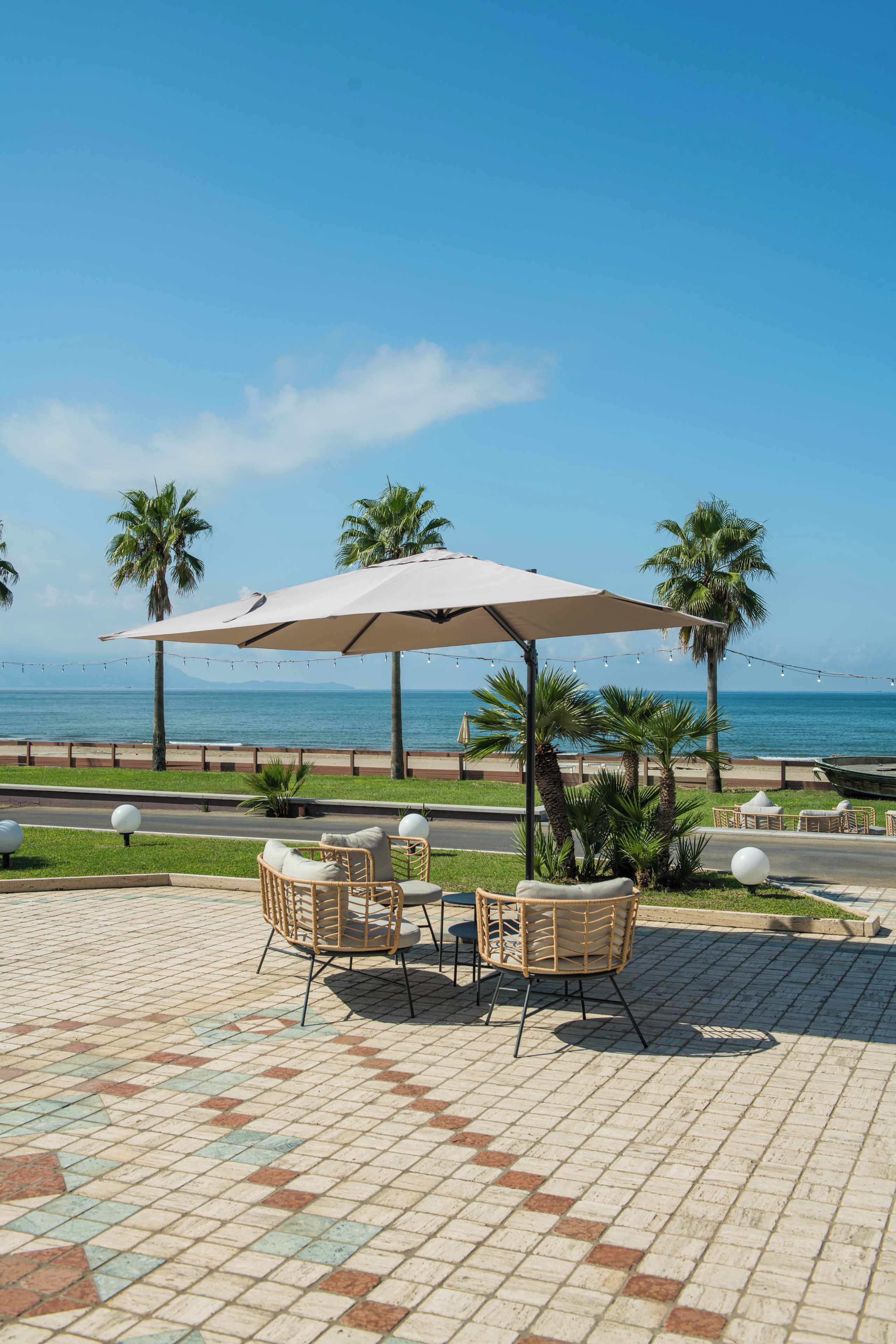 An outdoor seating area with a table and chairs under a canopy, overlooking a beach with palm trees and a clear sky.