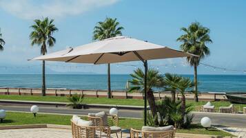 An outdoor seating area with a table and chairs under a canopy, overlooking a beach with palm trees and a clear sky.