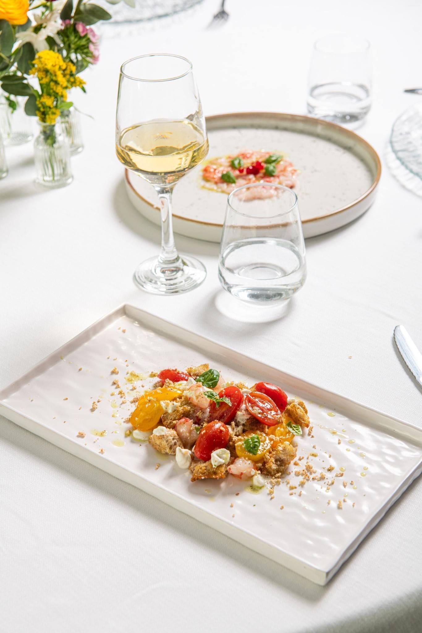 Une table à manger avec une assiette blanche remplie de nourriture, un verre de vin blanc et un vase de fleurs.