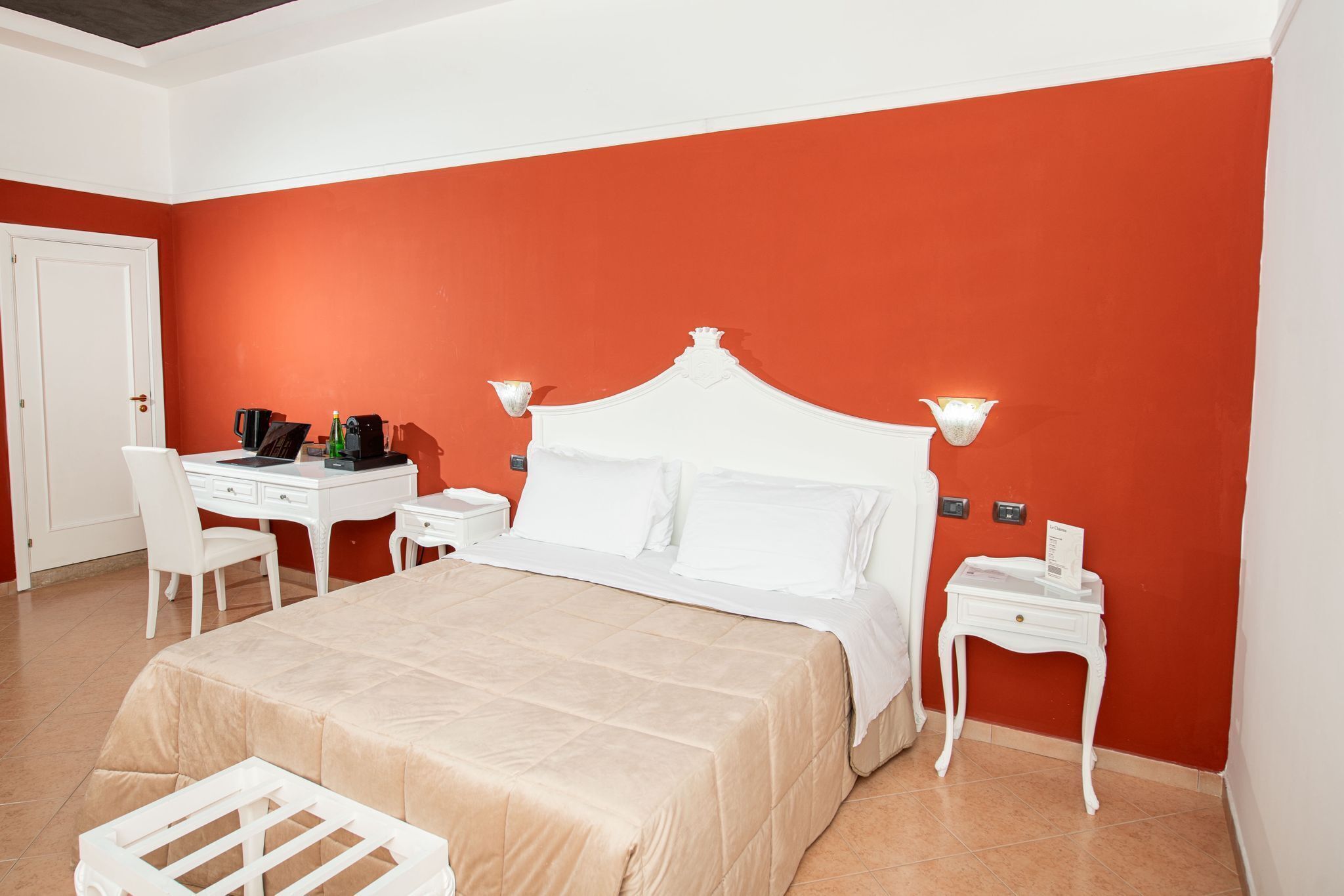 A hotel room with a bed, white desk, chair, bedside tables, and a red accent wall.