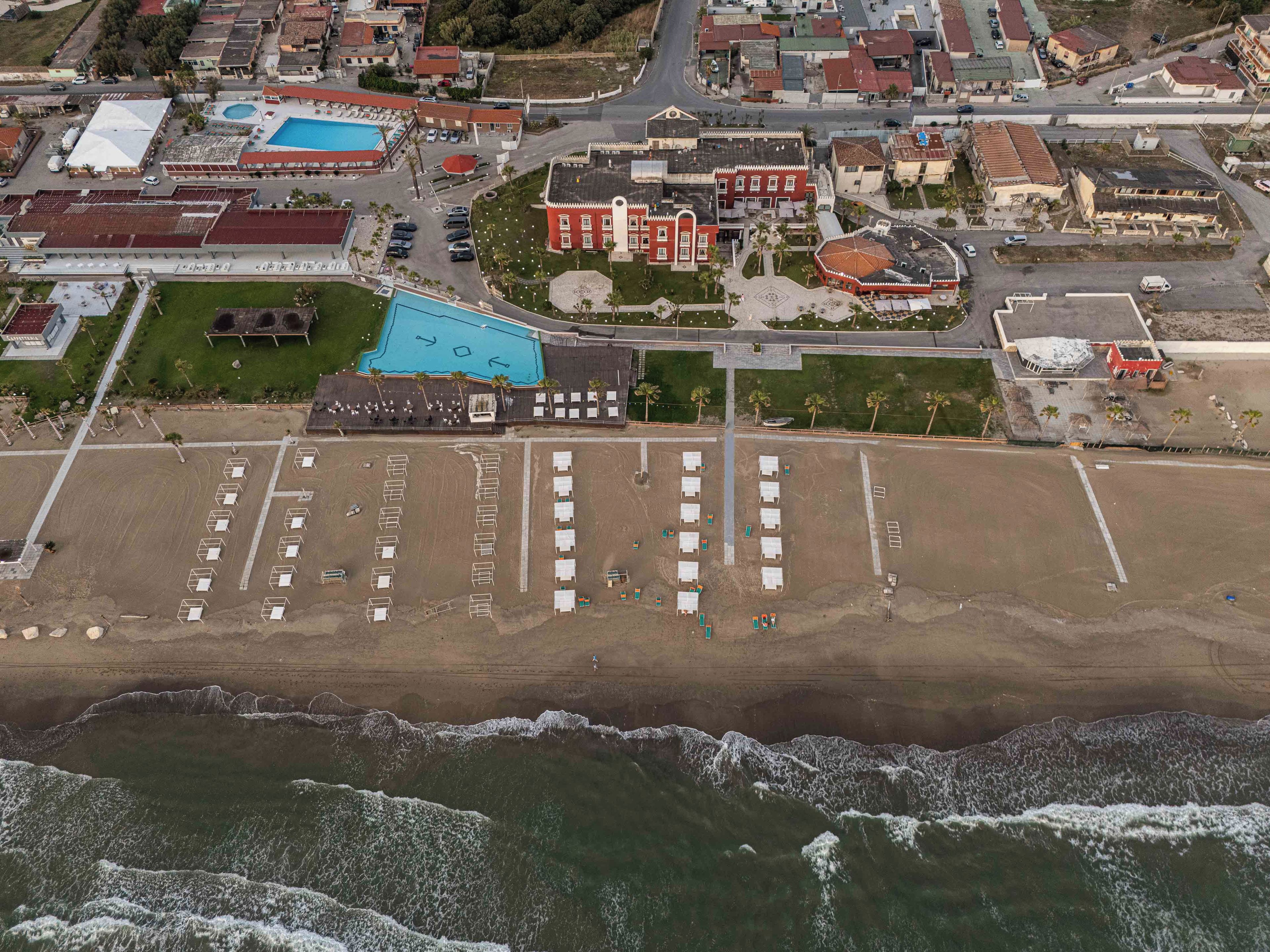 An aerial view of a coastal area with a beach, parking lot, and buildings including a hotel with a swimming pool.