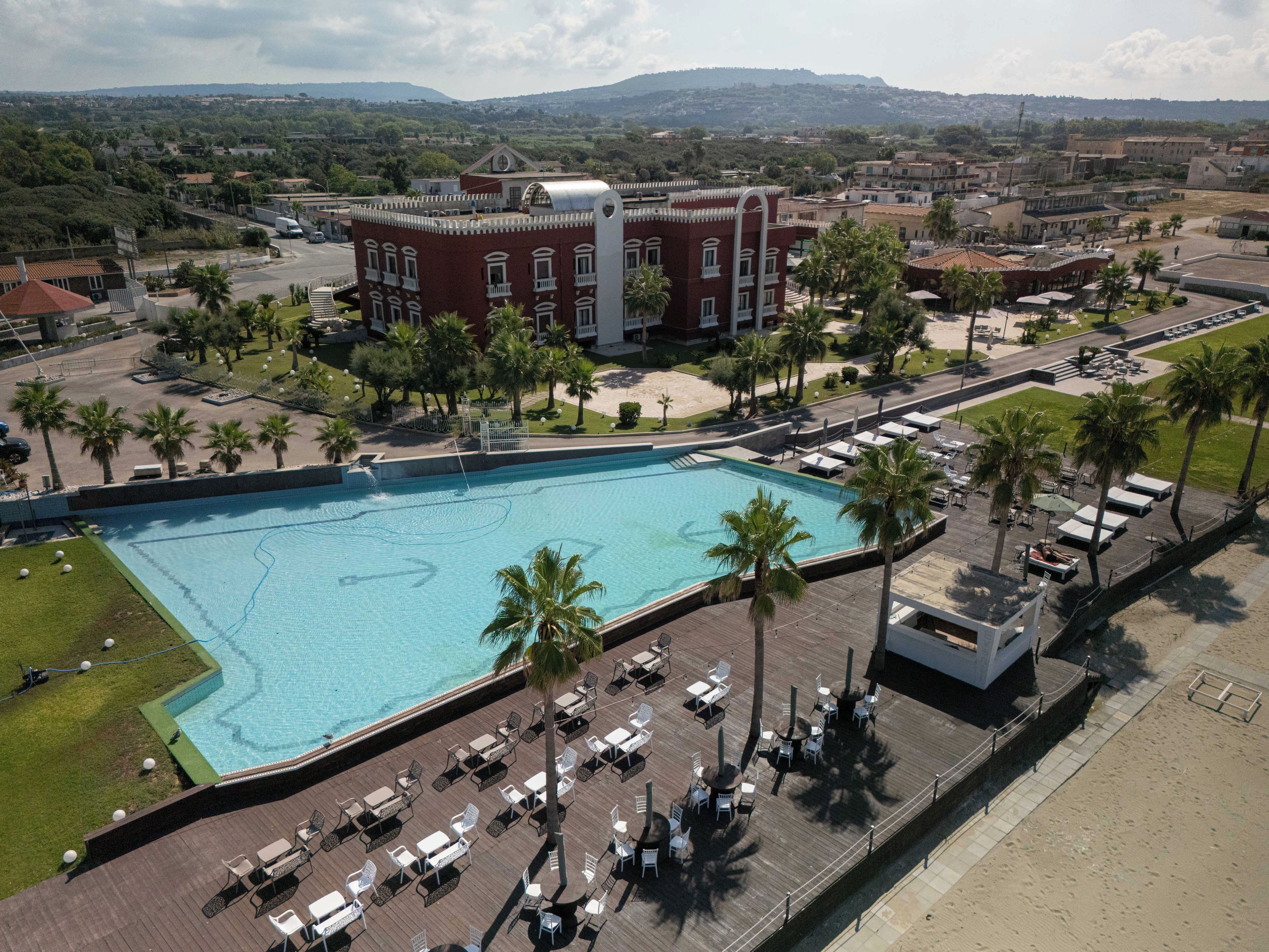 A large swimming pool with a deck area, surrounded by palm trees and a building with a distinctive roof.