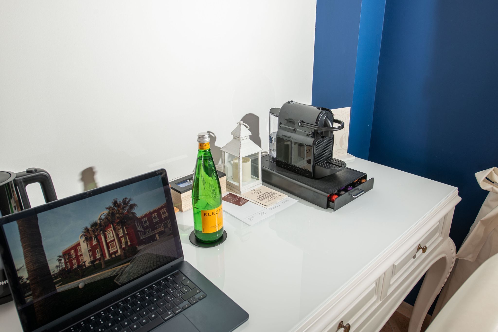 A laptop, a green bottle, a coffee machine, and a white desk in a room with a blue wall.