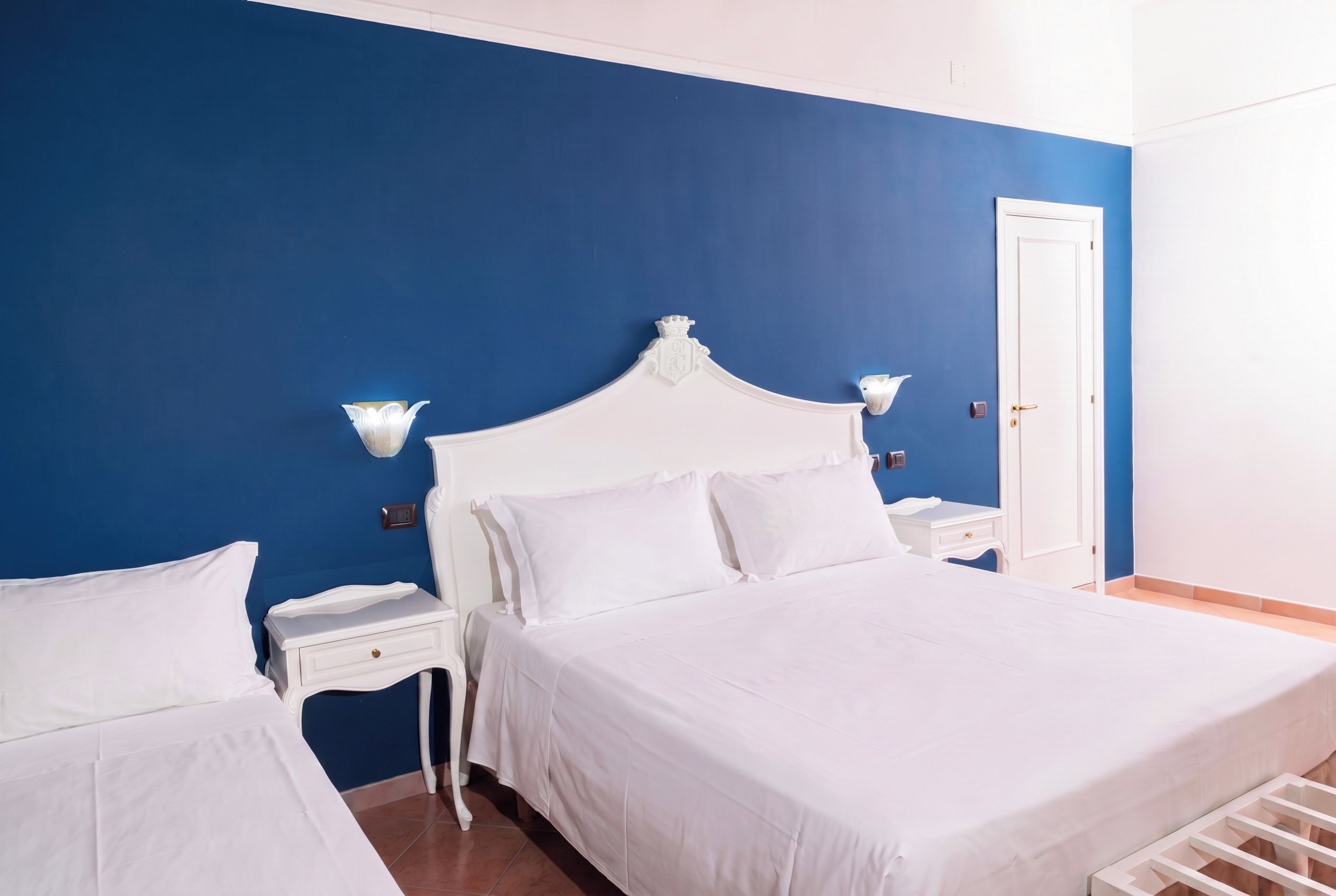 A hotel room with a large bed, a bedside table, a nightstand, a wall-mounted light, and a door.