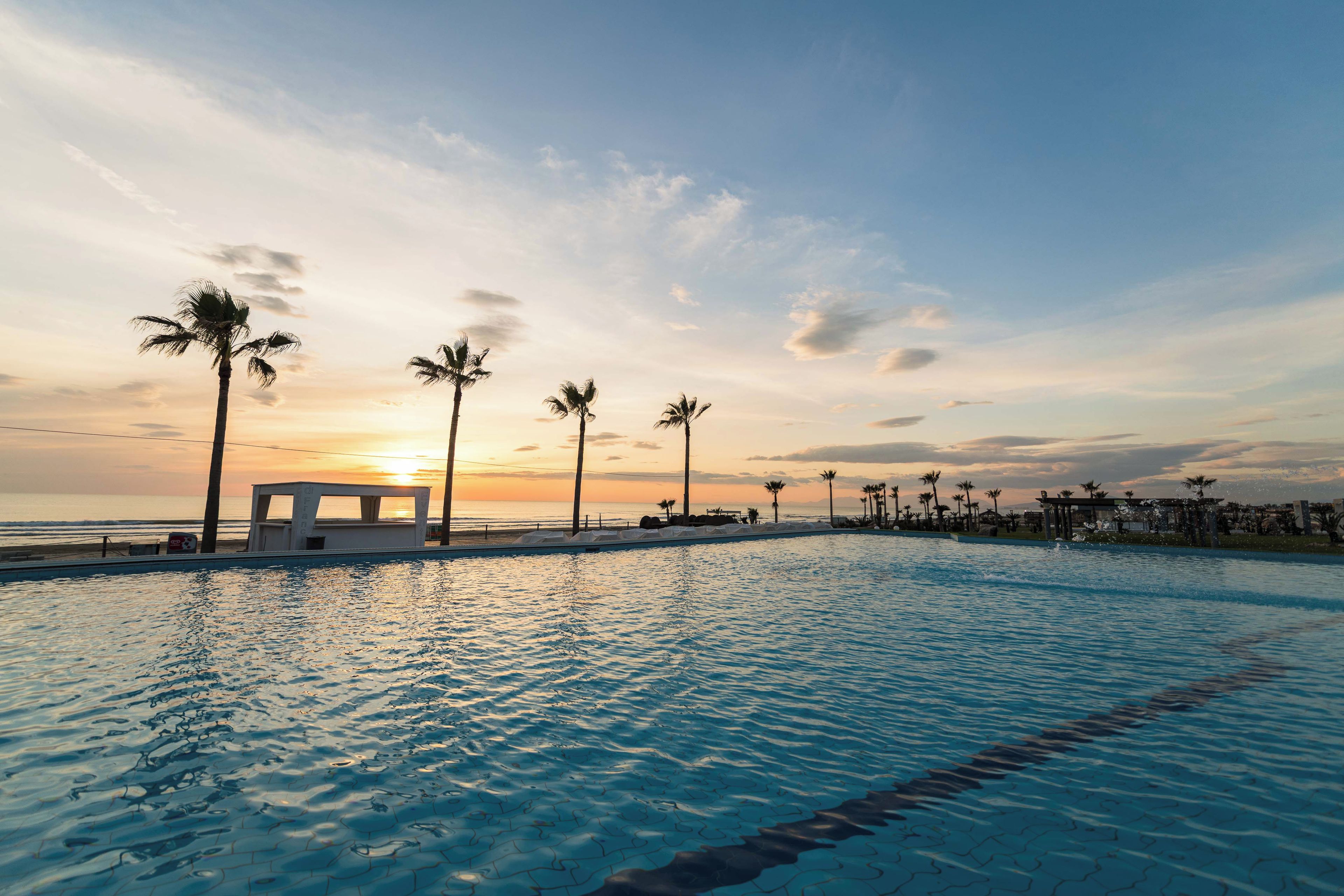 A swimming pool with a sunset view, palm trees, and a beach in the background.