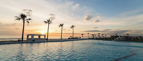 Une piscine avec vue sur le coucher de soleil, des palmiers et une plage en arrière-plan.