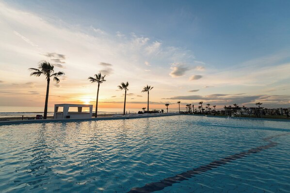 Una piscina con vista al atardecer, palmeras y una playa al fondo.