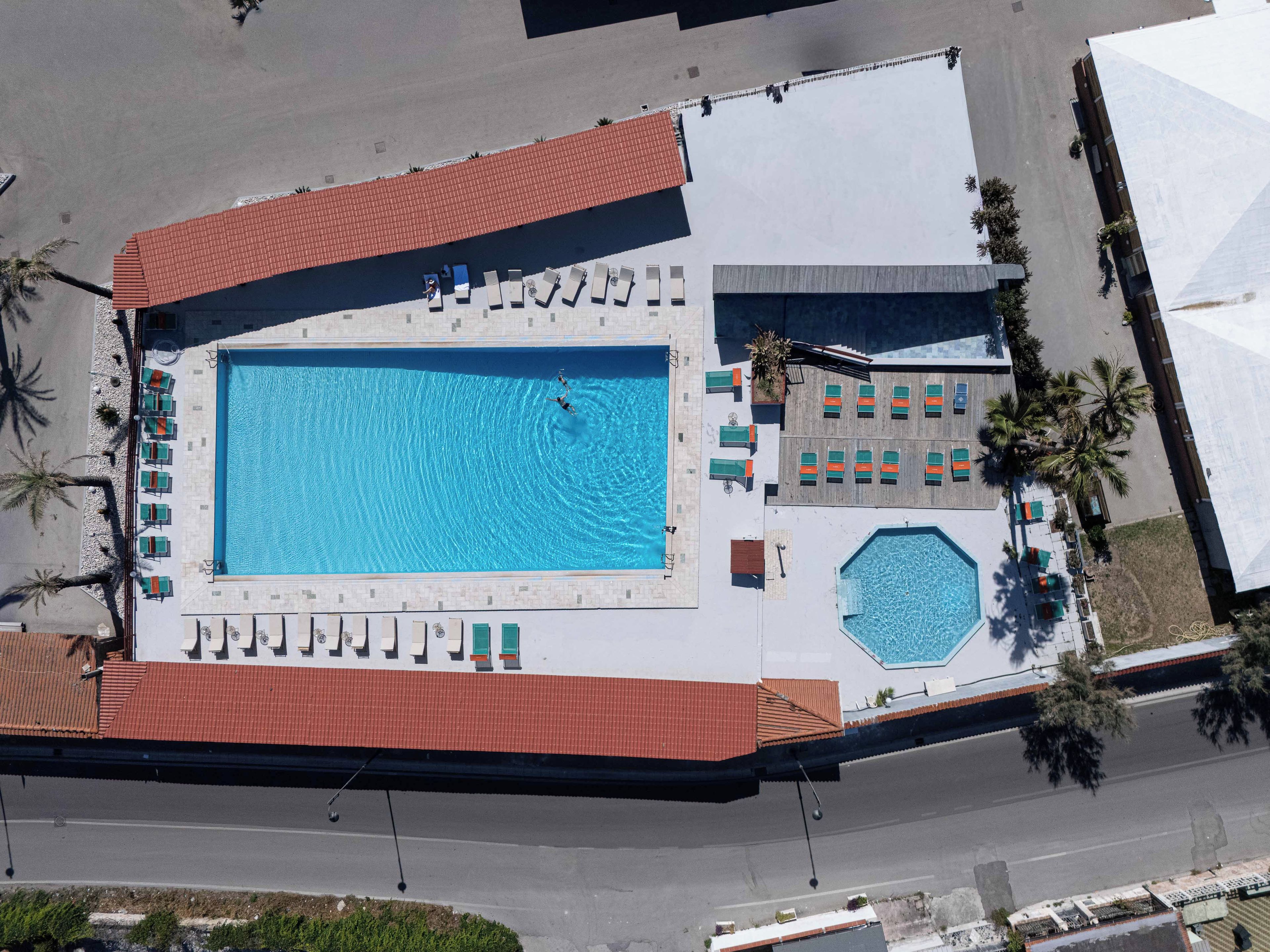 An aerial view of a hotel with a large rectangular swimming pool, multiple lounge areas, and a covered outdoor seating area.