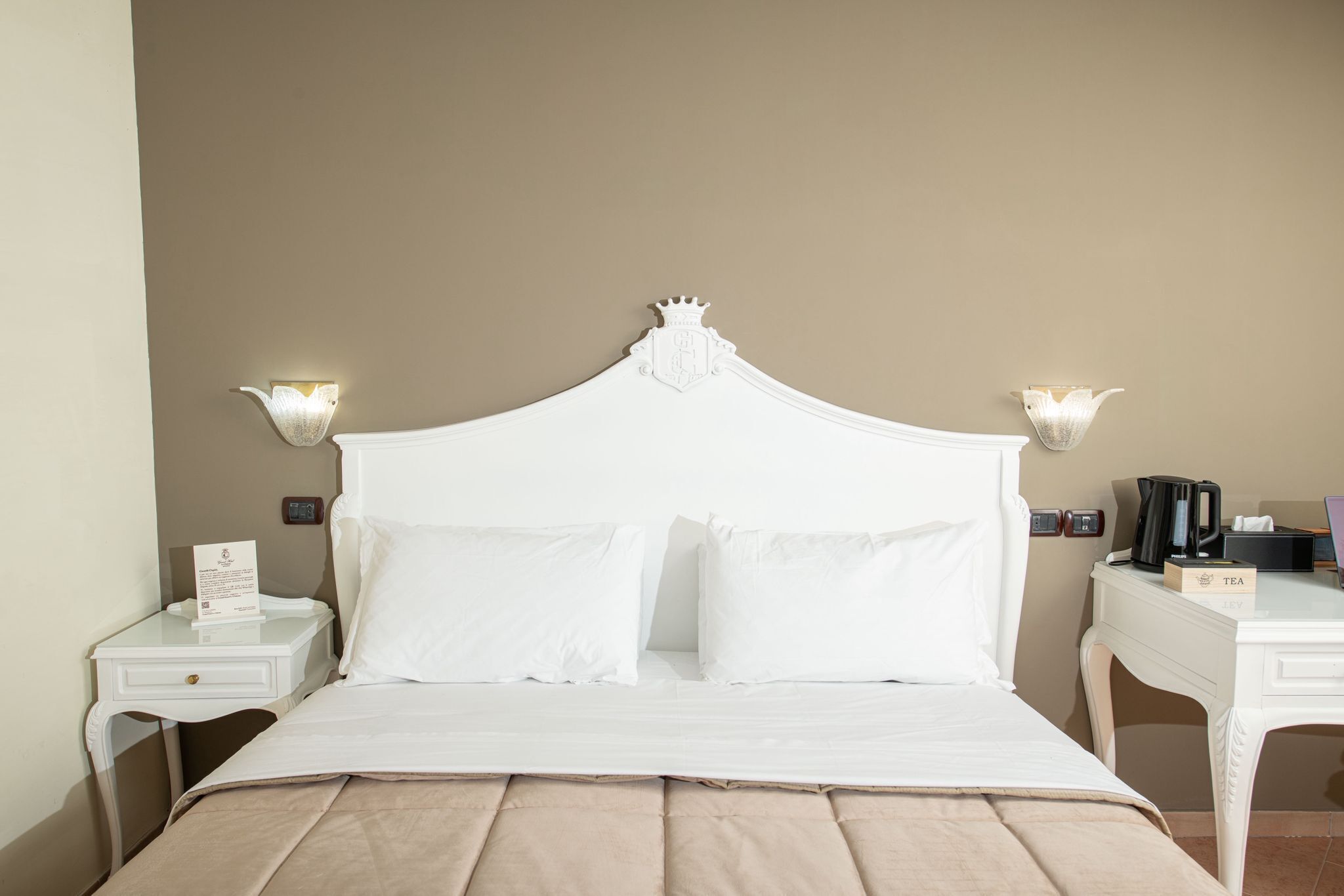 A neatly made bed with a white headboard, two bedside tables with lamps, a bedside table with a coffee maker, and a small table with a tea tray.